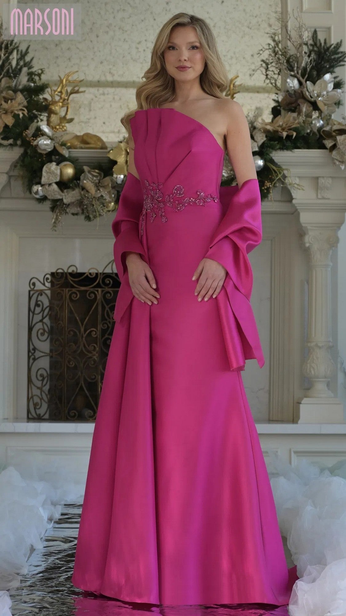 A woman poses indoors in front of a decorated fireplace, wearing the Marsoni MV1356 long formal dress in fuchsia with floral embellishments and a matching shawl.